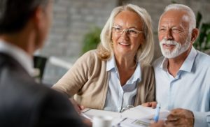 An older couple sits at a desk smiling and talking with a professional, who is partially visible in the foreground. The couple holds papers, suggesting a consultation or meeting in a bright, modern office.