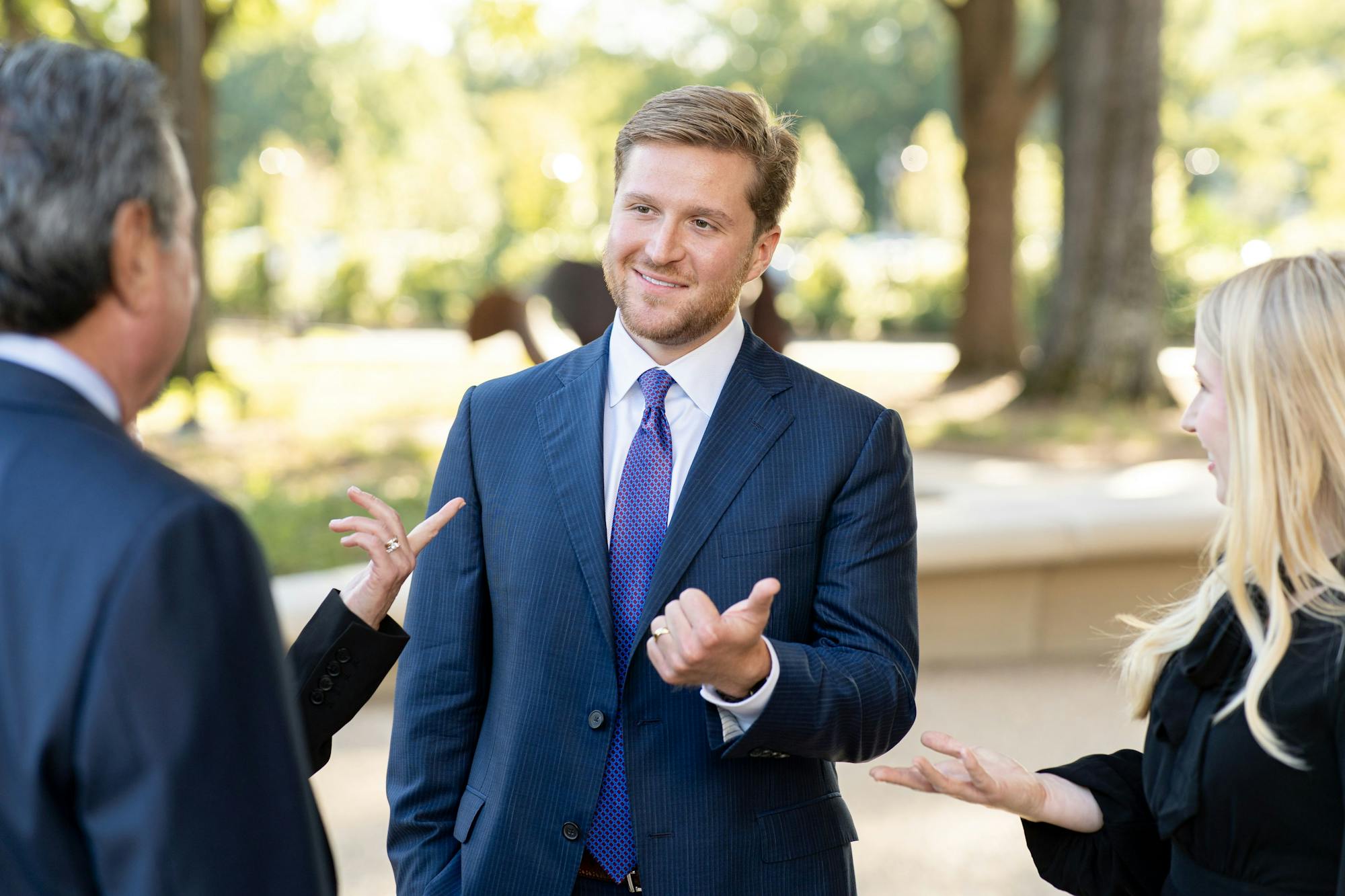 A man in a blue suit and purple tie smiles and gestures while talking with two people outdoors, surrounded by trees.