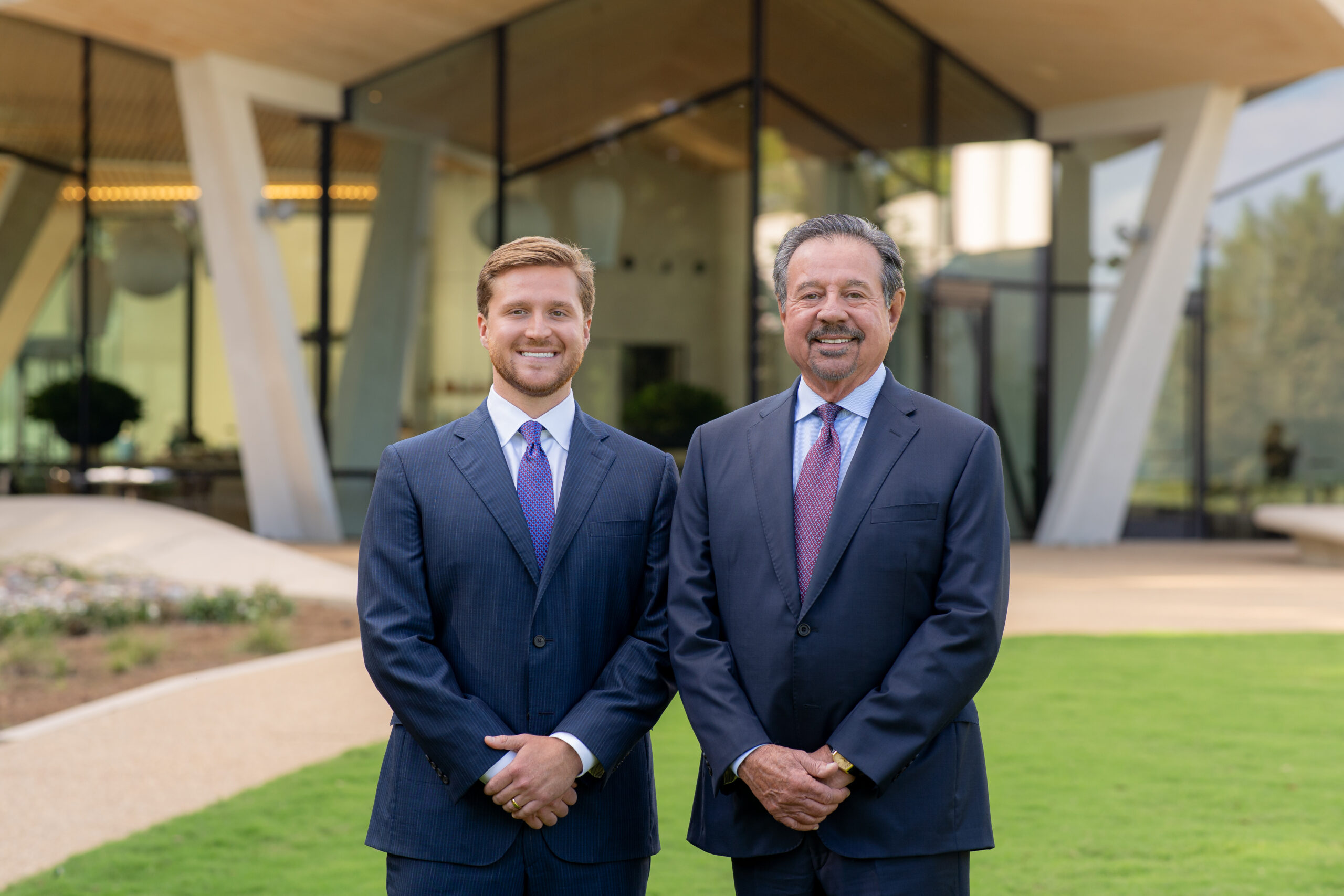 Two men in suits stand side by side, smiling, in front of a modern glass building with white pillars and a green lawn.