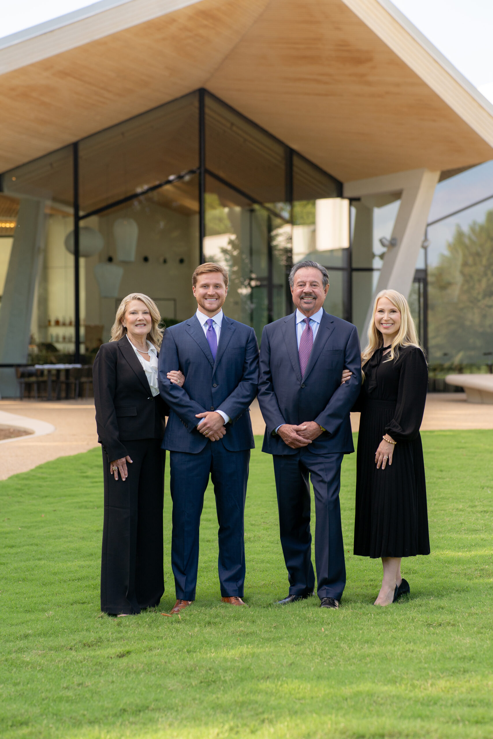 Four people, two men in suits and two women in formal attire, stand smiling on green grass in front of a modern glass building with a wooden roof.