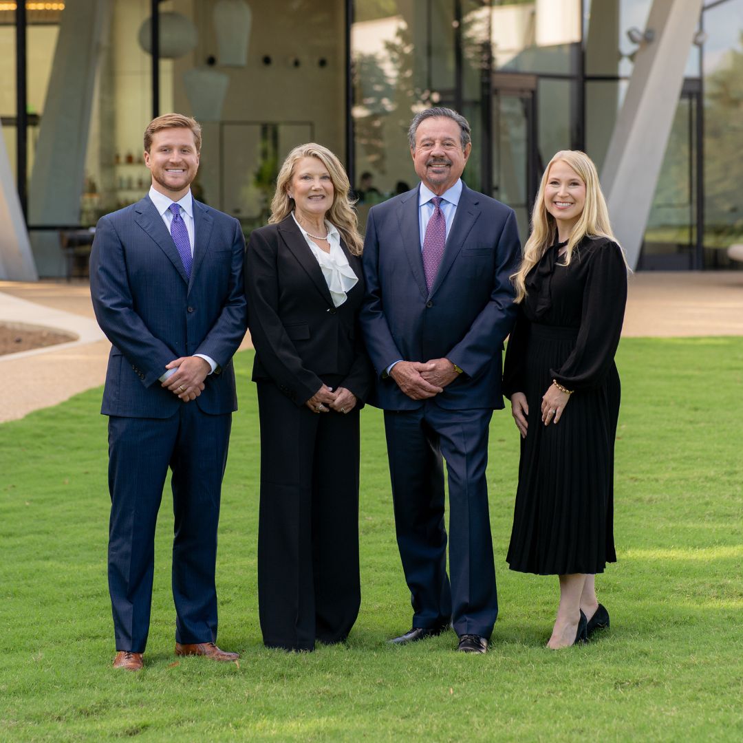 Four people in business attire stand on grass in front of a modern building, smiling and posing for a group photo.