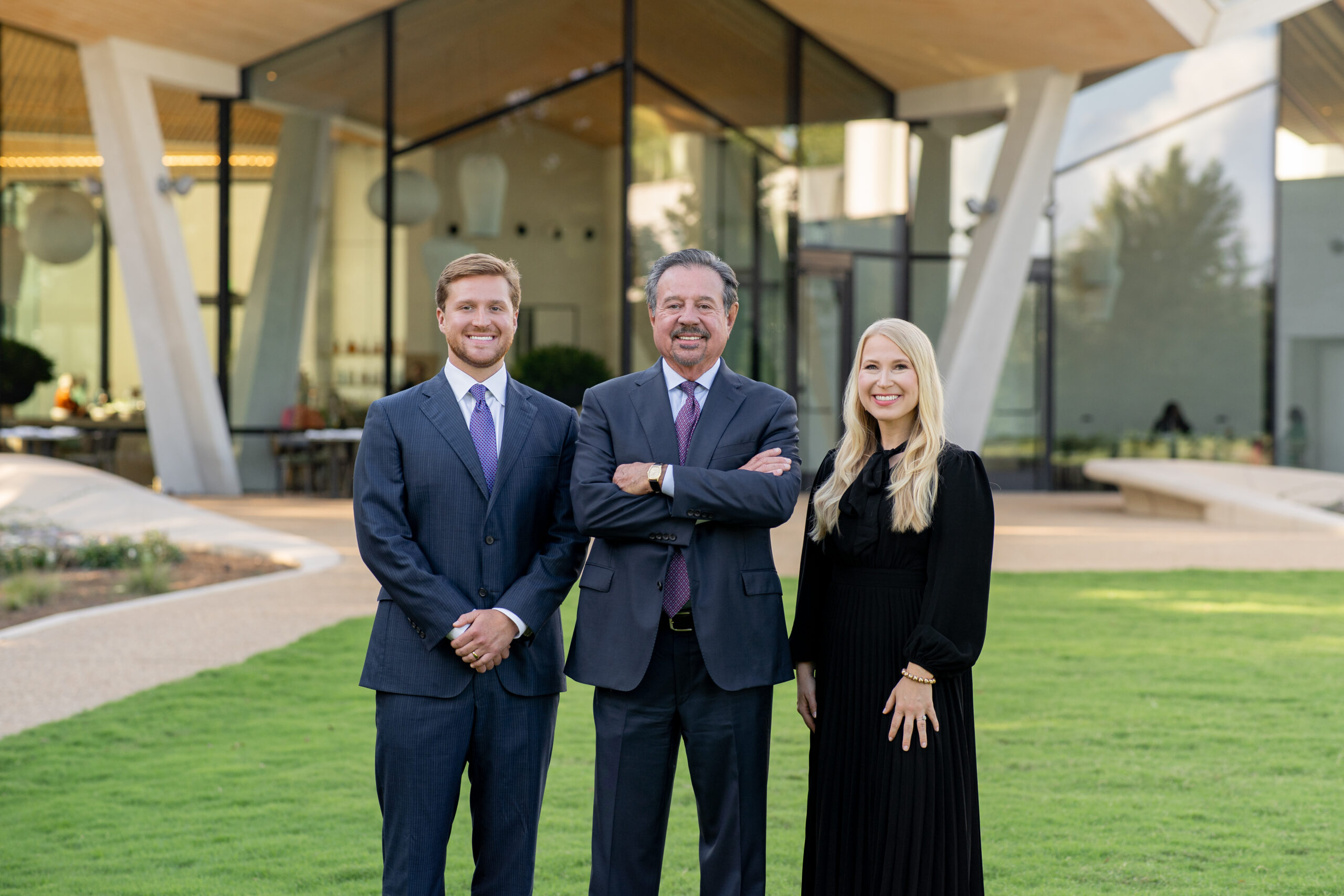 Three people in business attire stand smiling on grass in front of a modern glass building with geometric architectural features.